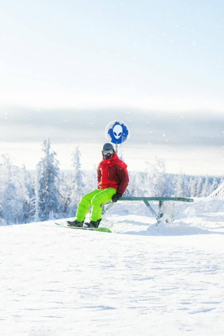 A snowboarder in vibrant gear sitting on a snowy bench with a winter landscape view.