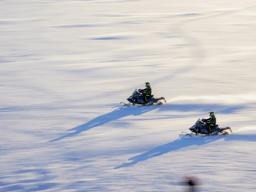 Two snowmobiles zoom across a vast snowy field in Lapland, Finland, during daylight.